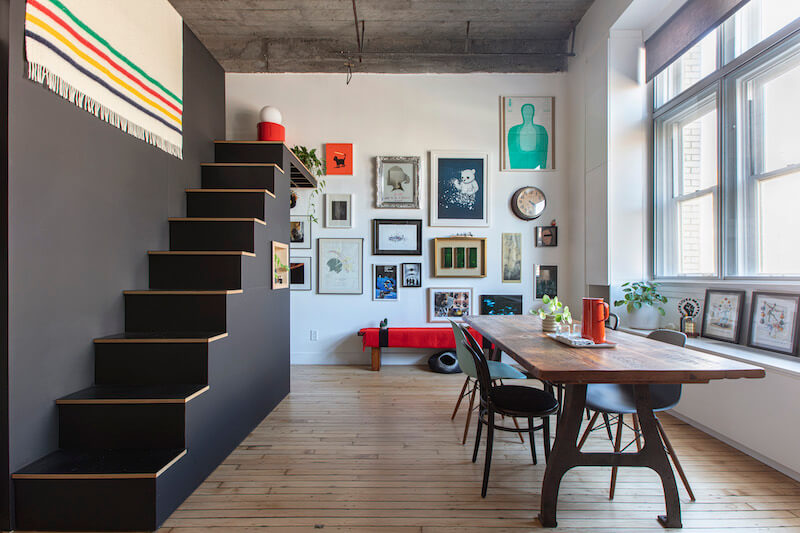 Loft dining area with gallery wall, black-painted mezzanine stair, and exposed concrete ceiling
