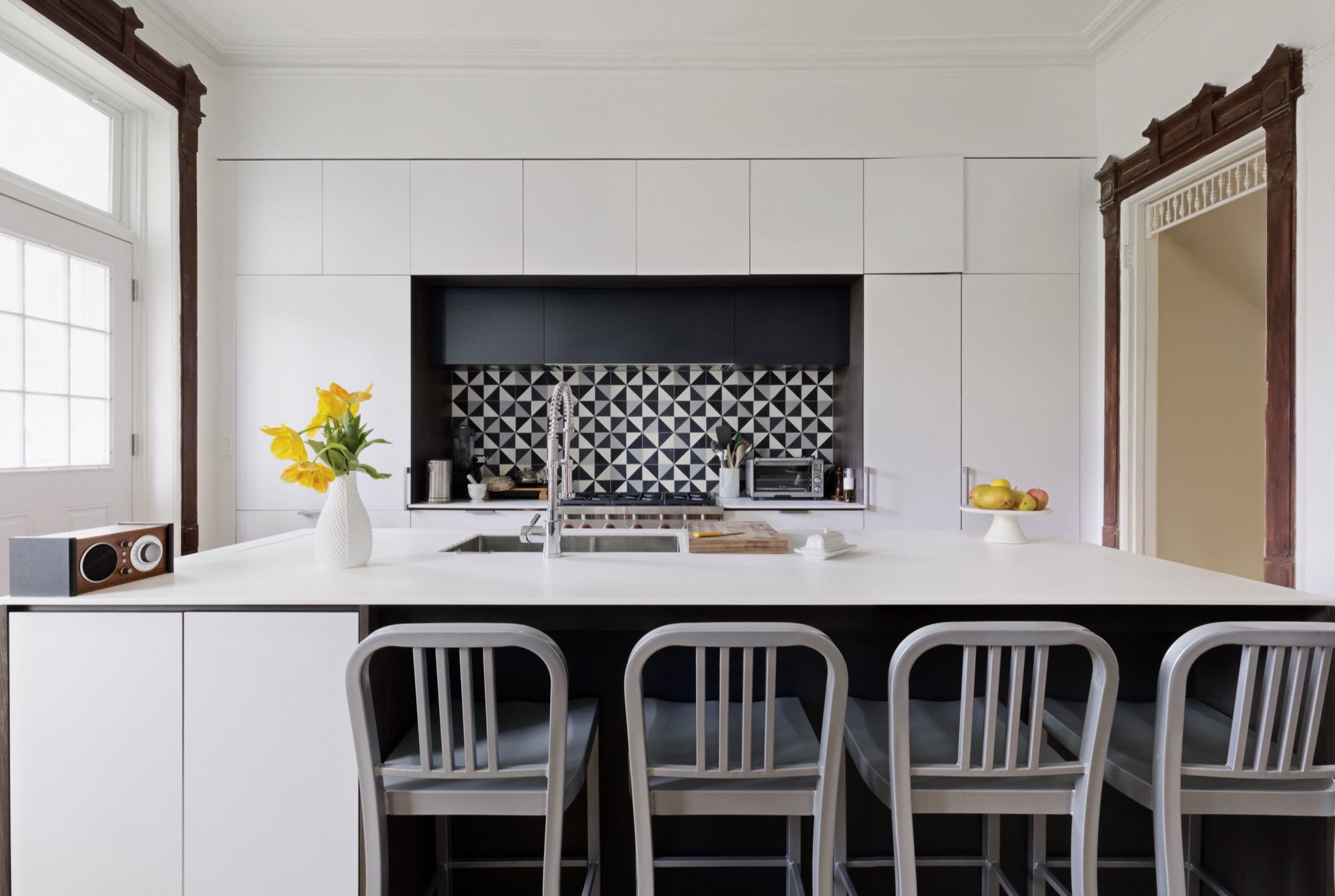 Proteus-renovated kitchen in a Brooklyn townhouse: white flat-front cabinets with a graphic black-and-white tile backsplash framed by preserved period moldings